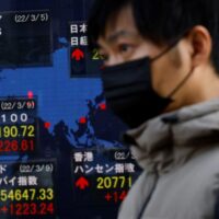 Ein Mann mit einer Schutzmaske geht am 10. März 2022 vor einer Maklerfirma in Tokio, Japan, an einer elektronischen Tafel vorbei, auf der die Aktienindizes verschiedener Länder angezeigt werden